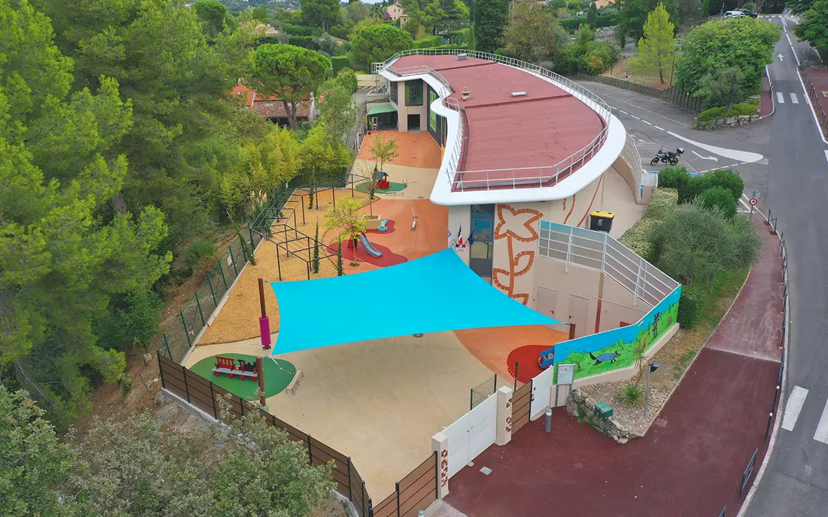 Une aire de jeux moderne avec toboggans, entourée d'arbres, près d'un bâtiment incurvé et routes. Couleurs vives et structures ludiques pour enfants.