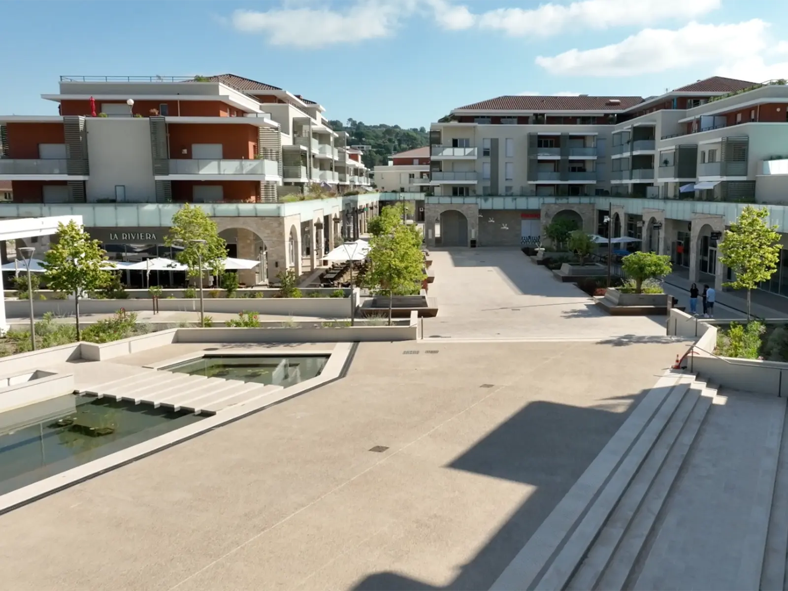 Une place moderne avec bâtiments résidentiels, commerces, et étang. Des personnes se promènent, profitant du cadre urbain et verdoyant, sous un ciel ensoleillé.