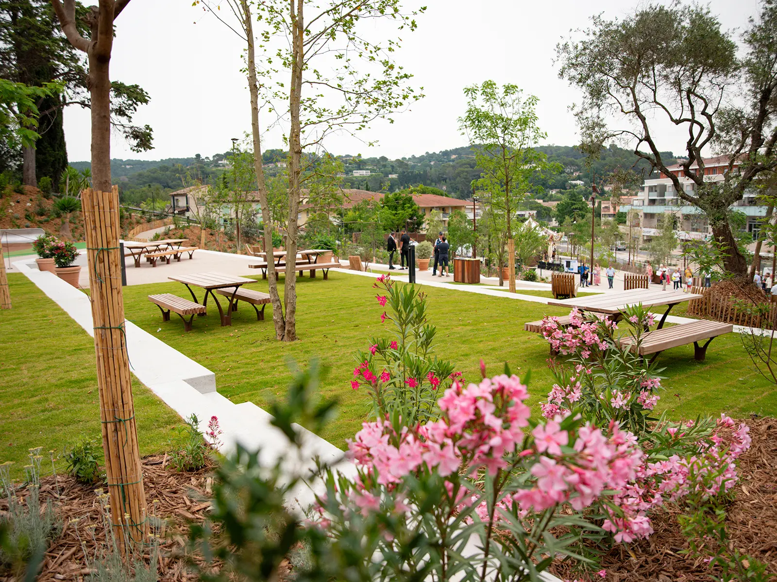 Pelouse avec tables de pique-nique, fleurs roses et arbres. Personnes se baladent dans le parc, entouré de végétation et de bâtiments modernes en arrière-plan.