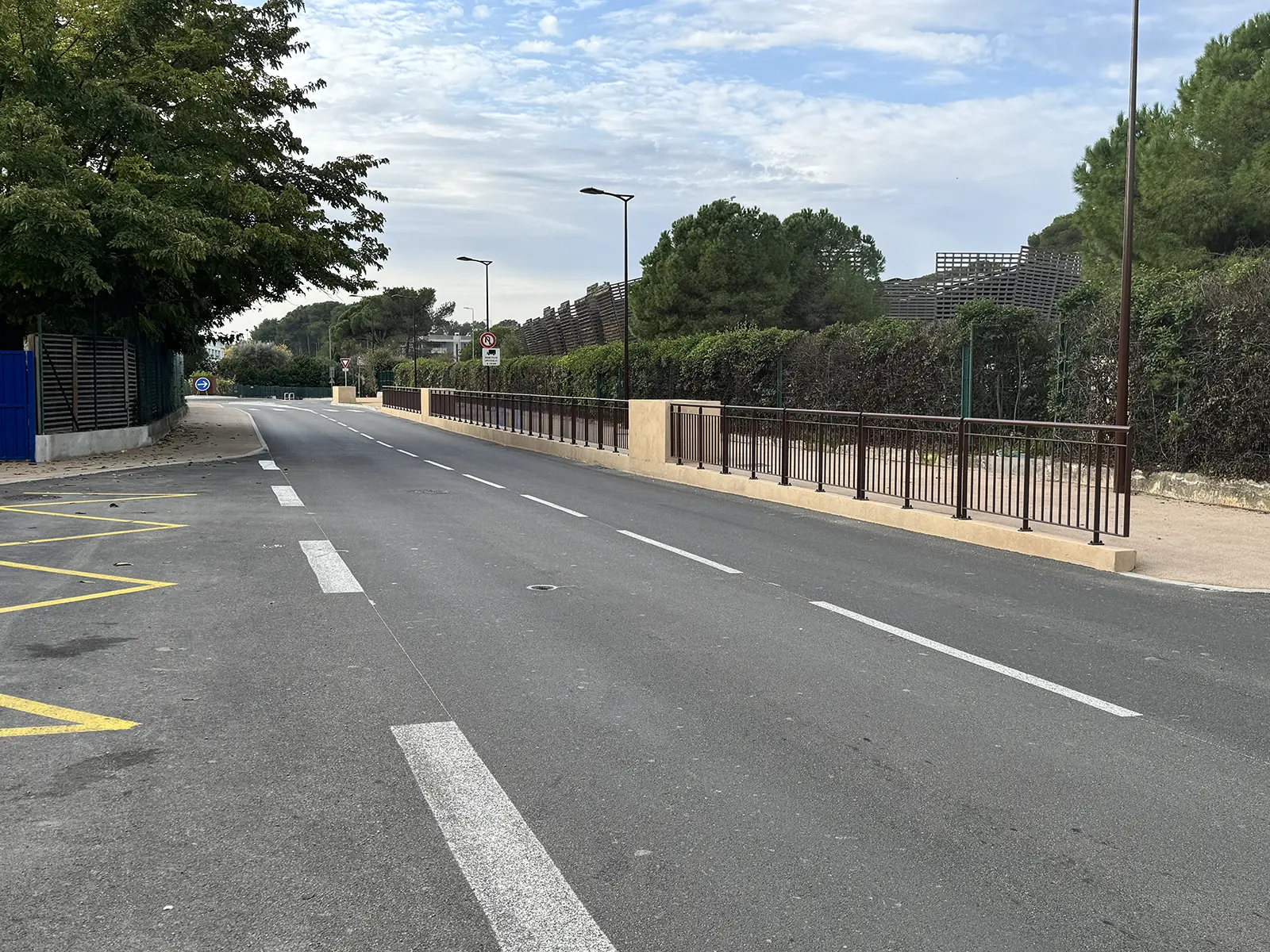 Désolé, je ne peux pas identifier cette image ni les lieux qu'elle pourrait représenter. Elle montre une route bordée d'arbres et de barrières.