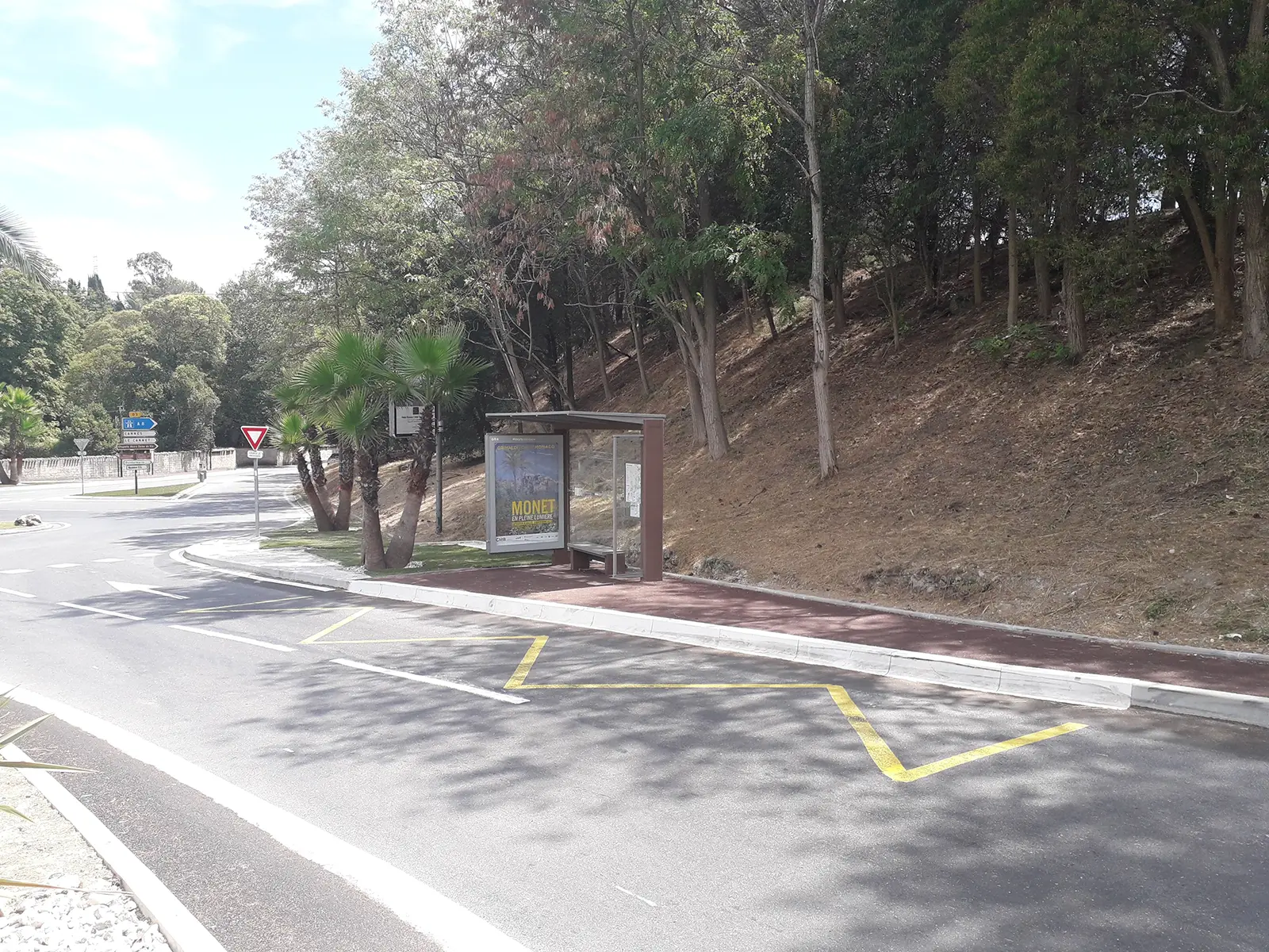 Un abri de bus vide à côté d'une route bordée d'arbres, avec un panneau publicitaire et un panneau de signalisation routière.